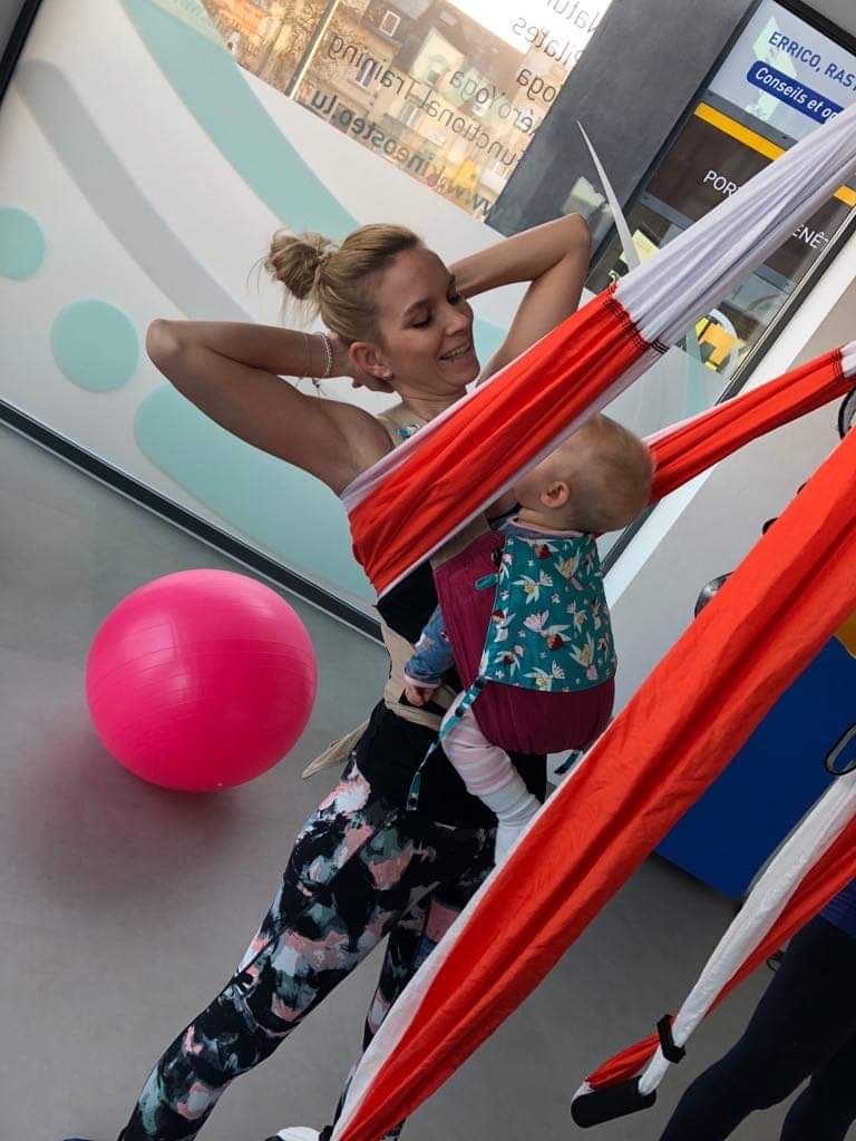 Mãe a alongar-se junto ao tecido, com o bebé ao peito num porta-bebés