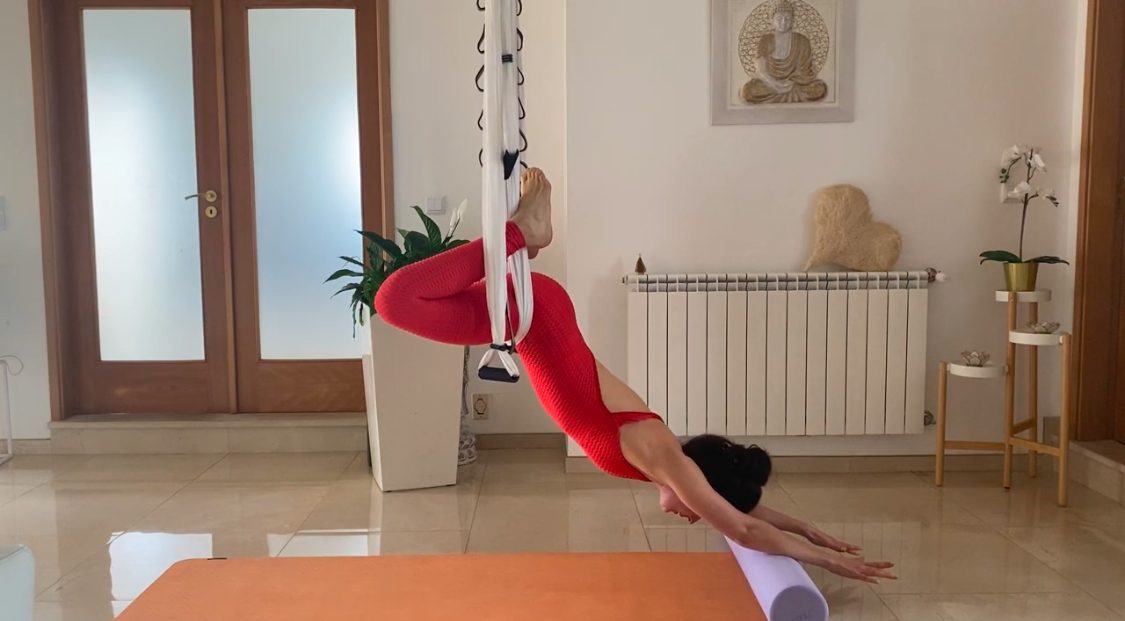 Prática de yoga aéreo em casa, com o tecido preso ao teto e um rolo no tapete